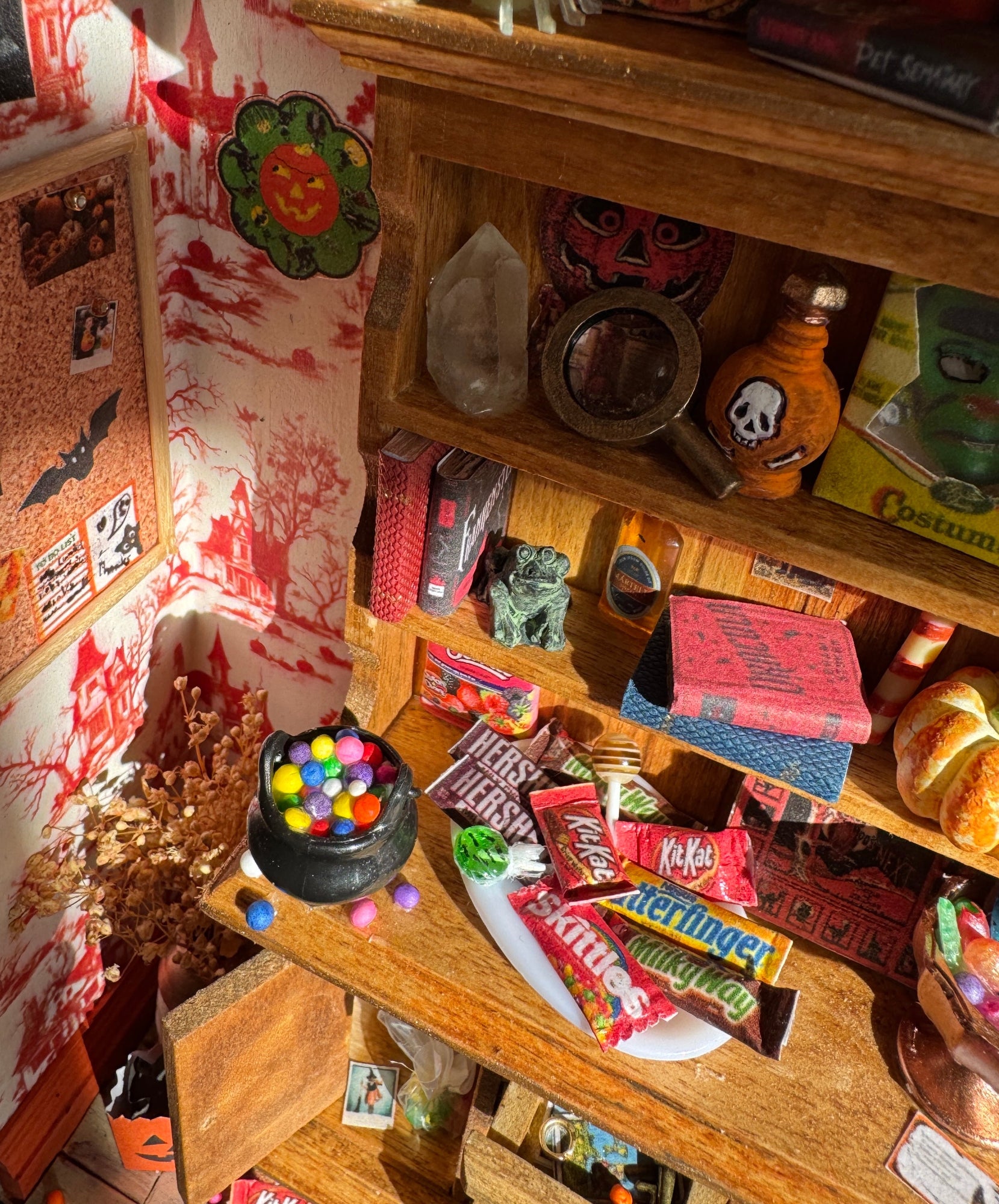 Close up of shelves at another angle showing crystals, potions, candy, horror books, a pumpkin, halloween mask, board games, and a magnifying glass. You can see loose candy on the floor, a jack o lantern trick or treat bag, dried yellow flowers. A cork board is hung on burnt orange and white haunted house wallpaper, with postcards and bats on it.