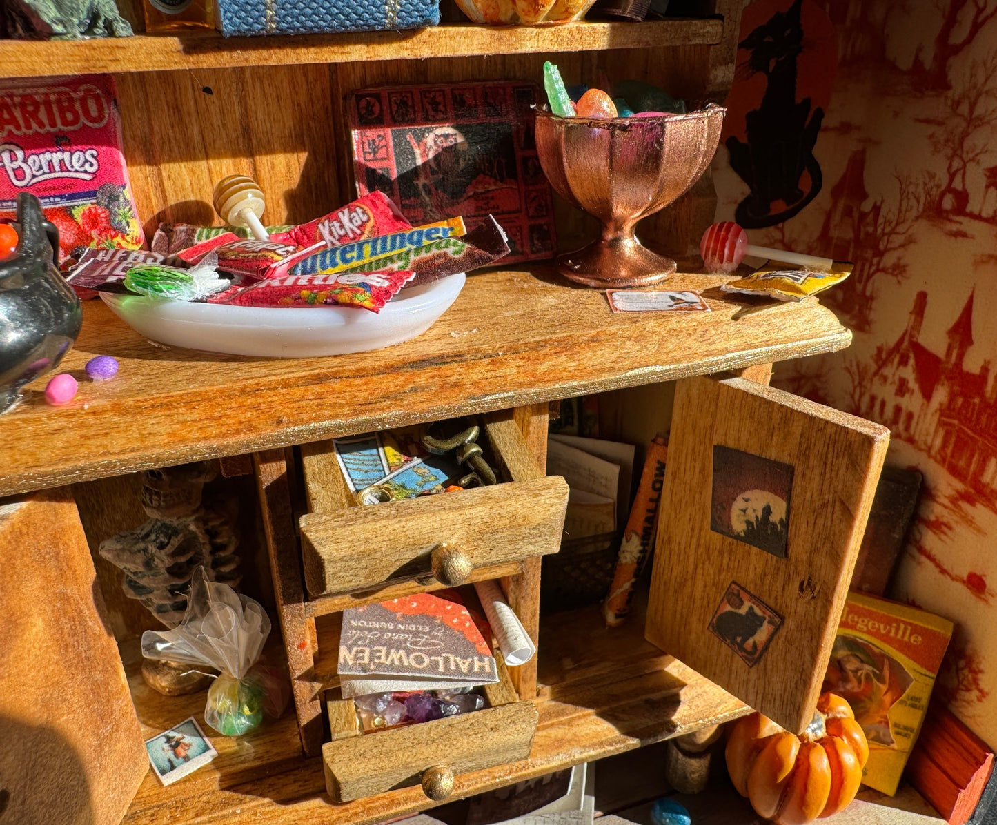 Another close up shot of the hutch with shelves full of candy bars, lollipops, loose jellybeans, and a copper bowl of candies. Open cabinet doors with bags of candy, photos, a skeleton bust, posters. Askew drawers with tarot cards, a key, rings, papers. A pumpkin and costume mask are on the floor leaned against a wall with haunted house wallpaper.
