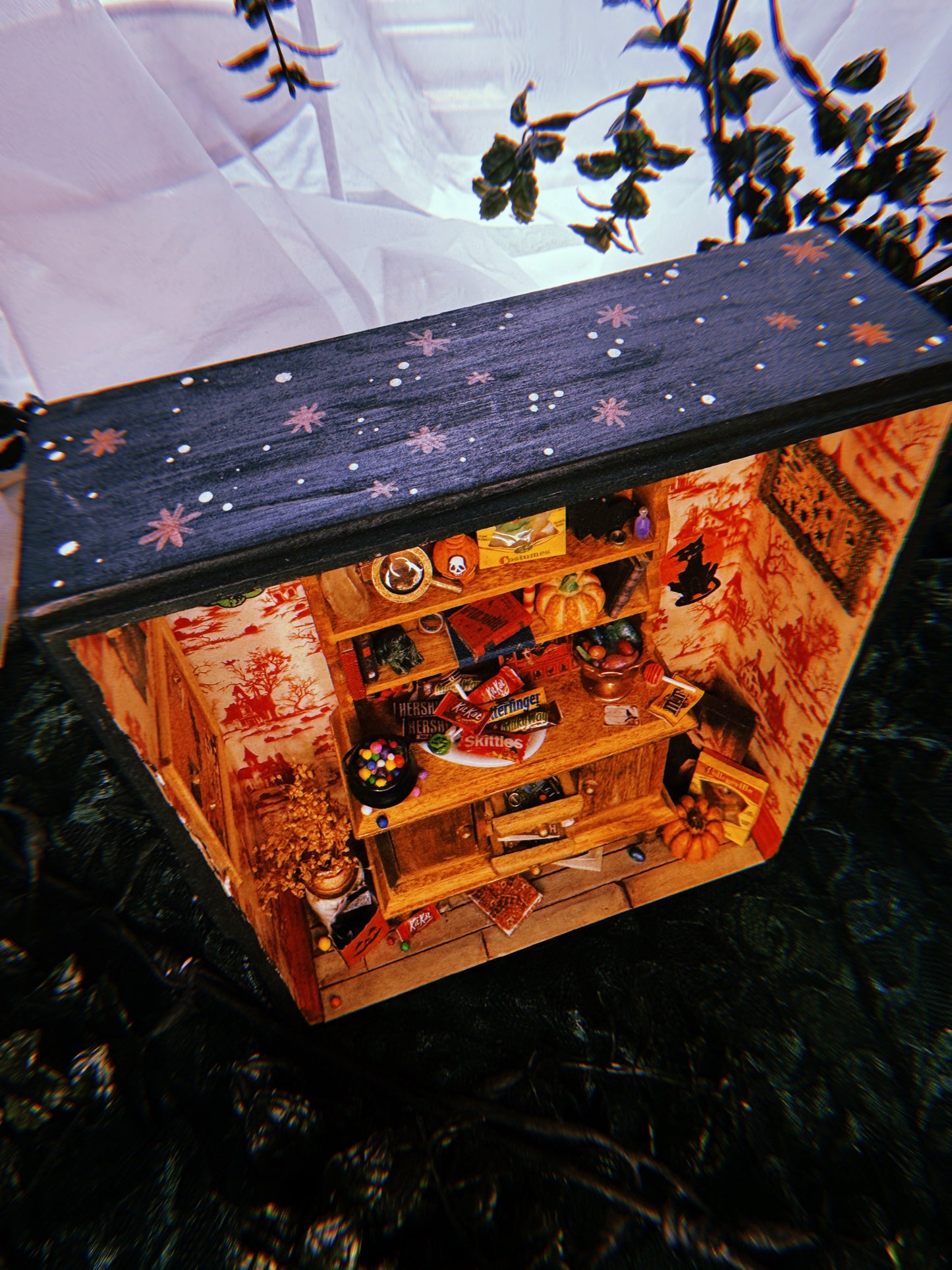 Another angle of the miniature Halloween-themed diorama that lets you see the inner details and painted outside. The view is from the top and shows the room full of festive candy, games, and spooky wall decorations. The outside is painted black with white speckles and burnt orange starbursts.