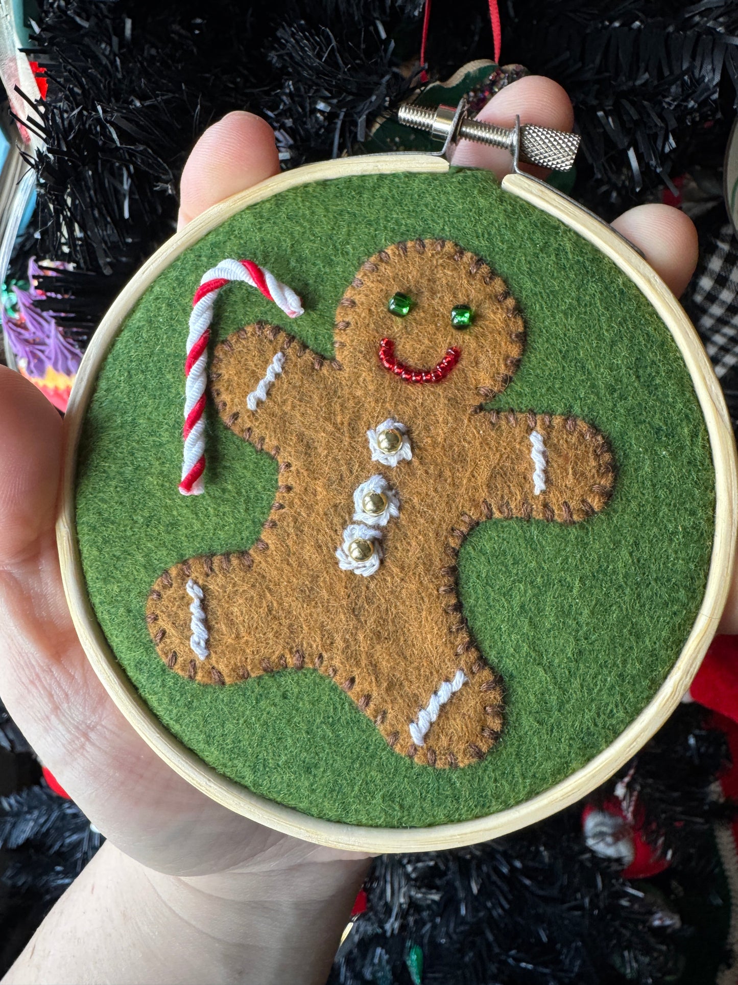 A hand holding a felt embroidery hoop with a gingerbread cookie design, featuring green frosting, white thread, and a candy cane.